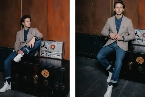 Junger Mann im lässigen Look: Sakko Herren sportlich zu Jeans, dazu weiße Sneaker. Ein Vintage-Tonbandgerät steht daneben.