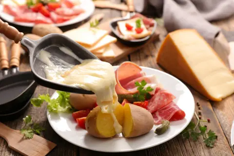 Roztopiony ser raclette spływa na ziemniaki i wędliny. Obok kawałek sera i talerz z dodatkami.