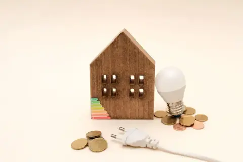 Drewniany domek z etykietą energetyczną, żarówką i monetami. Zastanawiasz się, ile prądu zużywa lampka nocna?