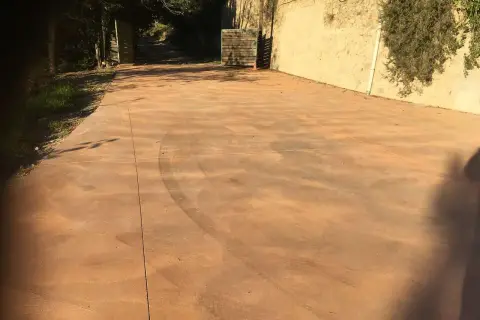 Un camino de **suelo hormigon pintado** de color terracota, con marcas de neumáticos, conduce a una puerta abierta.