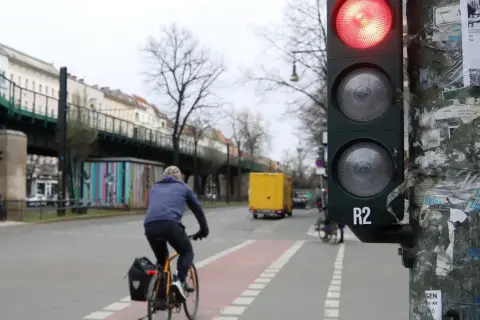 Ein Radfahrer wartet an einer roten Ampel. Im Hintergrund eine gelbe LKW und eine Hochbahn.