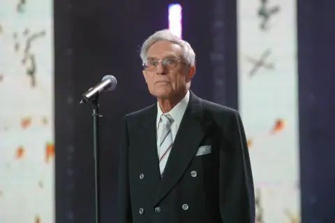 Starszy pan w okularach, Lucjan Szołajski, w czarnym garniturze i pasiastym krawacie stoi przy mikrofonie na tle sceny.