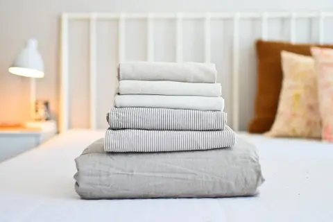 Neatly folded stack of clean bed sheets on a white bed, ready for how to wash bed sheets in washing machine.