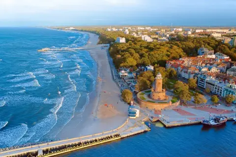 Widok z lotu ptaka na **latarnię morską Kołobrzeg** i wybrzeże Bałtyku. Widać plażę, molo i miasto.