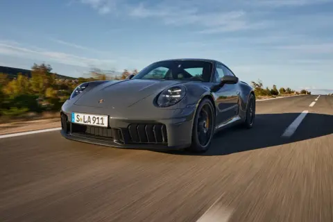 Szary Porsche 911 GTS sunie po krętej drodze, otoczony jesiennym krajobrazem.