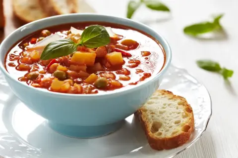 Aksamitna zupa solferino w błękitnej misce, udekorowana bazylią i parmezanem, obok kromka chleba.