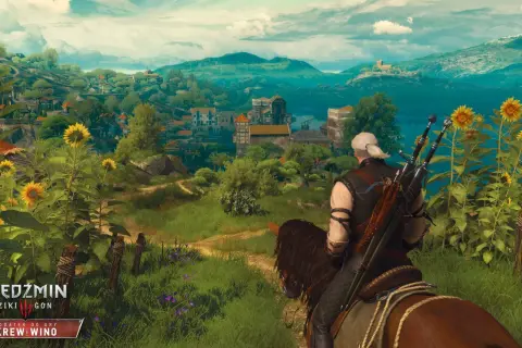 Geralt na koniu podziwia krajobraz z kwitnącymi słonecznikami, w oddali widać miasteczko. W tle góry i jezioro.