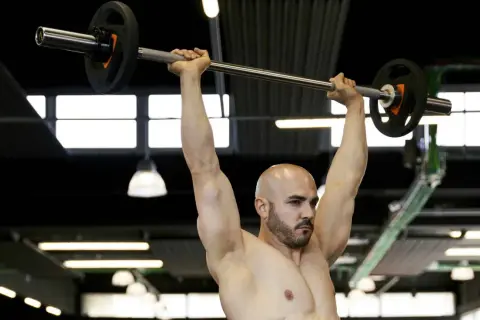 Uomo calvo con barba solleva bilanciere sopra la testa, concentrato sull'allenamento forza esplosiva.