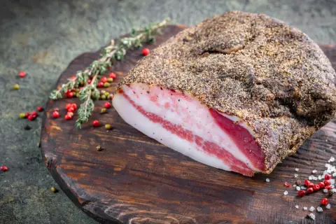 Pancetta co to? Wędzony kawałek boczku z widocznymi warstwami tłuszczu i mięsa, przyprawiony pieprzem, leży na drewnianej desce z ziołami i ziarnami pieprzu.