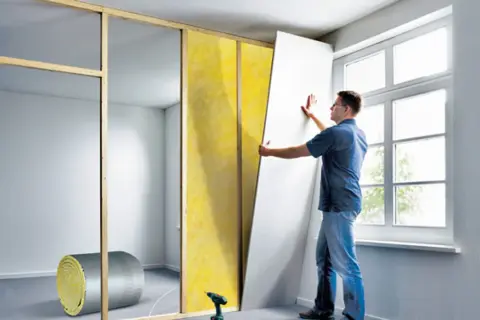 Mann zieht eine Gipskartonplatte an einer Holzlattung ein, um eine neue Wand zu bauen. Dämmmaterial liegt bereit.