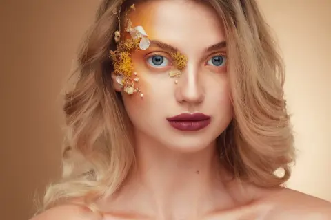 Fotografia beauty: kobieta z niebieskimi oczami, ozdobiona kwiatami i złotym makijażem na twarzy.