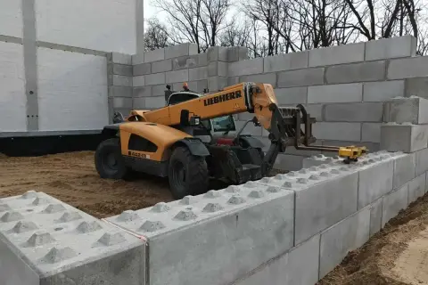 Żółty ładowacz teleskopowy Liebherr układa betonowe bloki, budując mur oporowy.