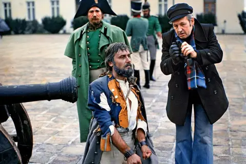 Scena z planu filmowego w polska lat 80. Uwięziony mężczyzna w zniszczonym stroju, obok żołnierze i reżyser.