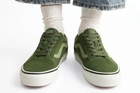 Zielone buty Vans na białym tle, widoczne od przodu, z nogawkami w białych skarpetkach i dżinsach.