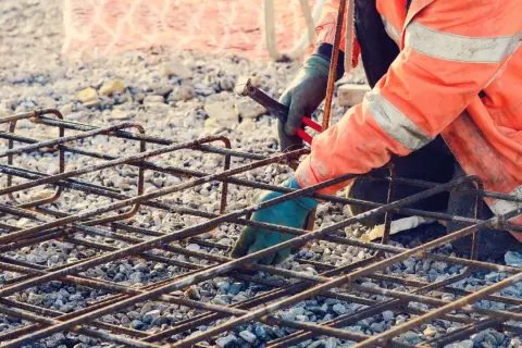 Budowlaniec w pomarańczowej kamizelce zbroi fundament, łącząc pręty. Zbrojenie rozproszone tworzy solidną siatkę.