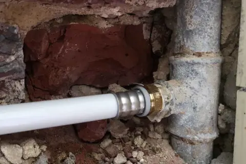 Un nouveau tuyau blanc est raccordé à un ancien tuyau de plomb pour l'eau. La maçonnerie est visible autour des canalisations.