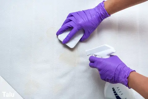 Mit lila Handschuhen wird erbrochenes aus Sofa entfernen hausmittel angewendet. Ein Schwamm tupft die Flecken, während eine Sprühflasche daneben steht.