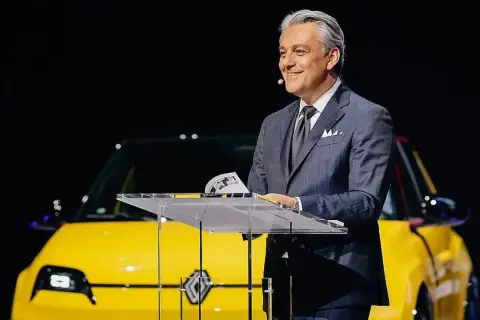 Hombre sonriente en traje, presentando un coche amarillo eléctrico, quizás un nuevo modelo de Volvo Renault.