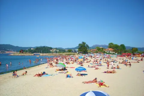 Día soleado en la playa Santa Marta de Baiona, con mucha gente disfrutando del mar y la arena bajo sombrillas.