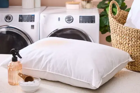 Learn how to get smell out of pillows with this guide. A stained pillow, spray bottle, and baking soda are shown, with laundry machines in the background.