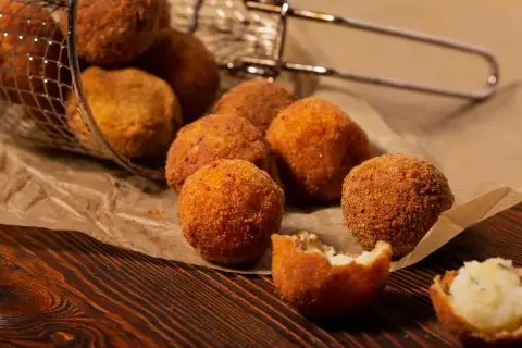 Croquetas de patata y pollo doradas y crujientes, algunas en una cesta metálica, otras sobre papel de estraza.