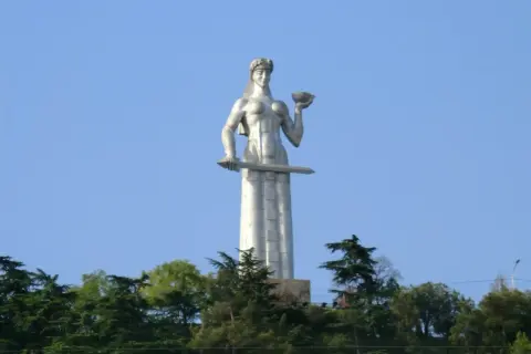 Matka Gruzja, symbol Tbilisi, trzyma miecz i puchar. Monumentalna postać wznosi się nad zielenią.