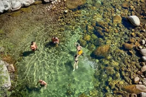 Gente se baña en un río cristalino con fondo de piedras, cerca de un **ponton de la oliva**.