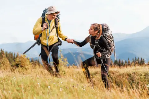 Coppia di escursionisti in montagna si aiutano a salire un pendio erboso, con zaini e bastoncini da trekking.