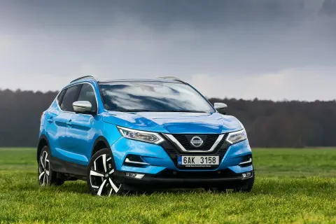 Niebieski Nissan Qashqai 1.6 benzyna na tle zielonej łąki i pochmurnego nieba.