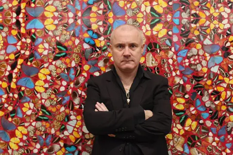 Damien Hirst stoi przed ścianą ozdobioną jego dziełami z motyli.