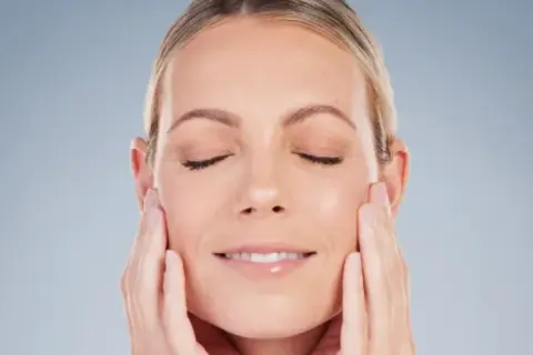 Mujer con ojos cerrados y una sonrisa suave, acariciando su rostro para realzar su óvalo facial.