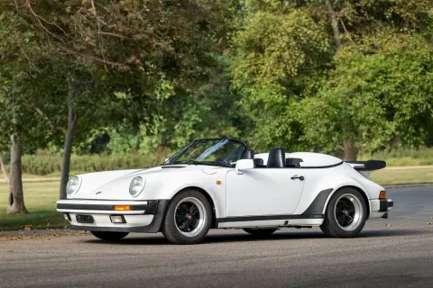 Biały Porsche 911 Speedster z lat 80. z czarnym dachem i spojlerem, zaparkowany na tle drzew.
