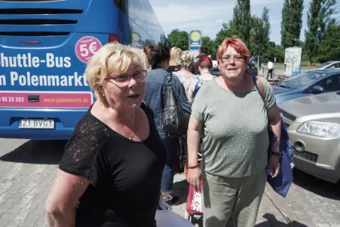 Zwei Frauen warten auf den Shuttle-Bus zum Polenmarkt. Der blaue Bus wirbt mit "5€ Fahrpreis".
