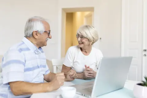 Couple âgé souriant devant un ordinateur, discutant de leur compte bancaire sous curatelle renforcée.