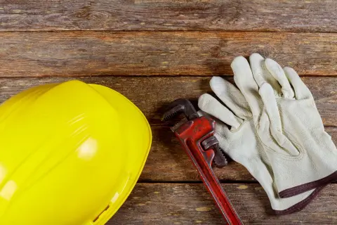 Żółty kask, klucz hydrauliczny i rękawice na drewnianym tle. Zastanawiasz się, dlaczego woda schodzi, a jest bulgotanie w rurach? Czas na fachowca!