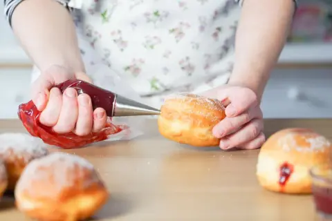 Hände füllen mit Marmelade gefüllte, zuckerbestäubte **bolas de fraile** mit einem Spritzbeutel.