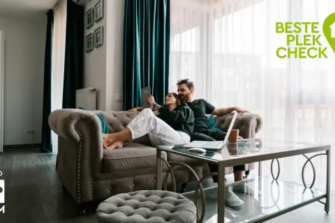Een stel overweegt wel of niet verhuizen, zittend op de bank met een laptop. De "Beste Plek Check" logo suggereert hulp bij deze beslissing.