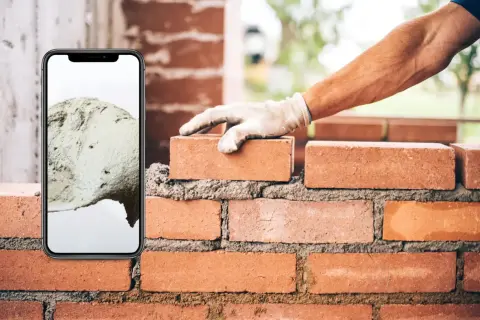 Teléfono móvil muestra cemento, mano con guante coloca ladrillo en pared.