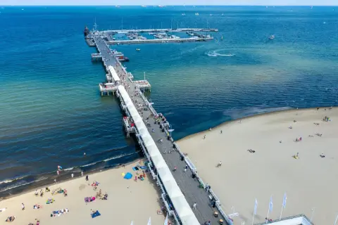 Słoneczny dzień nad Bałtykiem. Molo w Polsce tętni życiem, spacerują po nim tłumy ludzi. Na plaży odpoczywają plażowicze, a w oddali widać jachty.