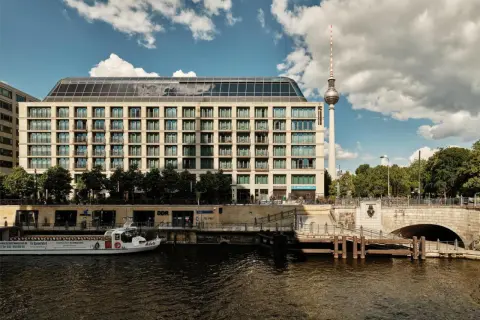 Nowoczesny budynek hotelu Radisson Blu Berlin nad rzeką Sprewą, z widoczną w tle wieżą telewizyjną.
