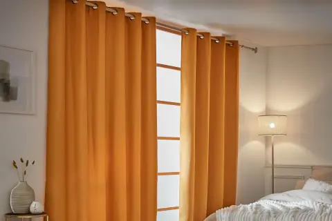 Cortinas aislantes naranjas cubren una ventana, creando un ambiente acogedor. A la izquierda, un cuadro abstracto y un jarrón con espigas.