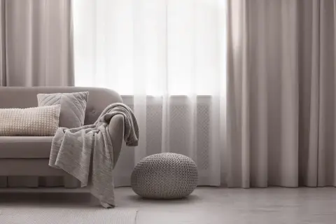 Elegancka dekoracja okien w salonie: szare zasłony i firanki tworzą przytulną atmosferę. Sofa z poduszkami i pledem zaprasza do relaksu.