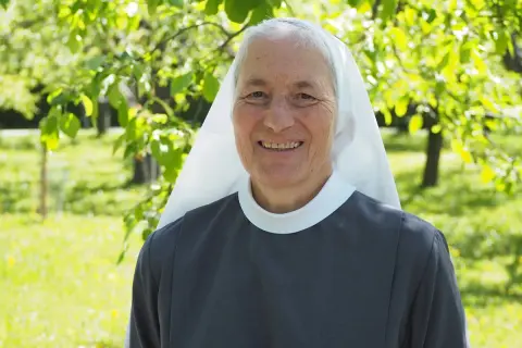 Lächelnde Nonne im Garten. Sie hat graue Haare und trägt eine graue Ordenskleidung mit weißem Schleier. Das Leben ist nicht fair, aber sie strahlt Hoffnung aus.
