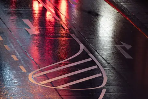 Nasse Straße mit roten und weißen Lichtreflexionen. Pfeile auf dem Asphalt zeigen die Fahrtrichtung an, wie bei einer anhalteweg formel.