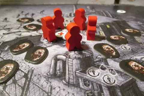 Czerwone pionki graczy walczą z zombie na planszy gry. W tle widać zniszczone budynki i barykady. To gra planszowa o tematyce atak zombie.
