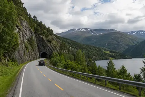 Svensk bil på väg mot en tunnel i Norge. Vacker bergig natur med sjö och moln.
