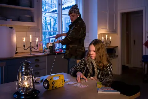 Två personer i ett kök med levande ljus och en sprit kamin. En flicka spelar kort vid ett bord, medan en person lagar mat vid spisen.