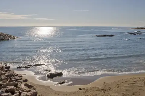 Costa de Villaricos con un faro en un rompeolas de rocas. El sol brilla sobre el mar azul y la arena dorada.