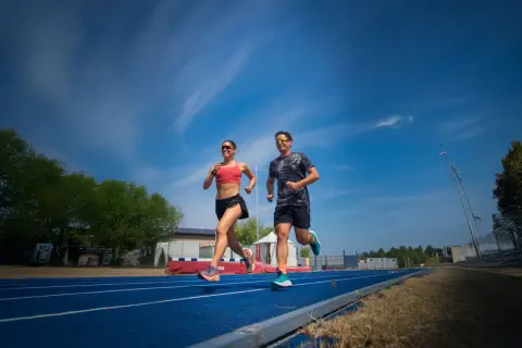 Coppia corre su pista blu, un allenamento intenso per la loro prossima corsa a tappe. Cielo azzurro e nuvole bianche.