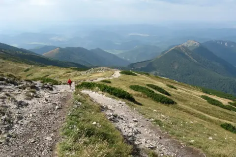 Wędrówka po górach Słowacji na weekend. Widok na zielone wzgórza i doliny, kamienista ścieżka, samotny turysta w czerwonej kurtce.
